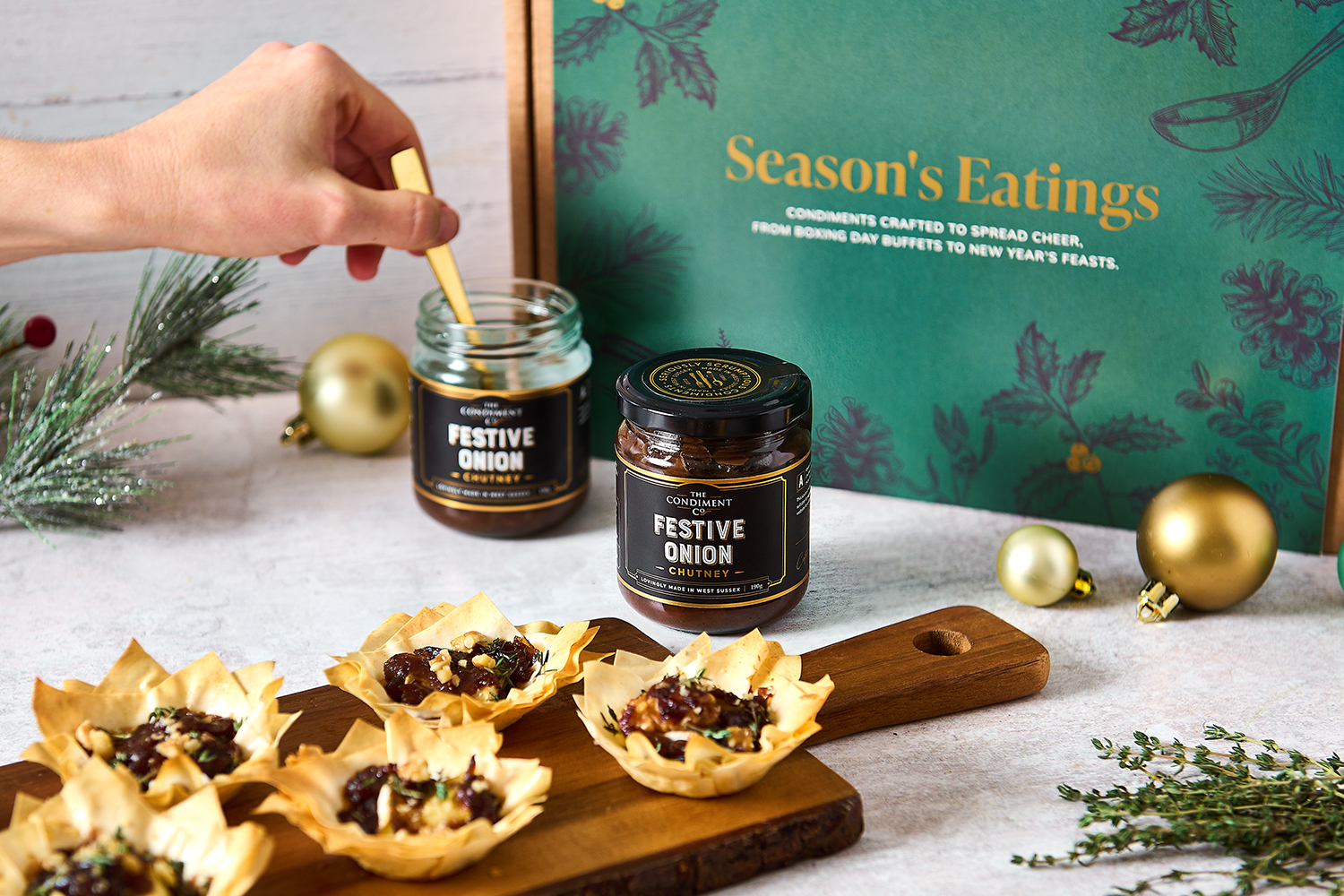 A wooden platter of pastry parcels filled with a dark chutney, in front of a hand dipping a spoon into an open jar and a green-labelled Condiment Co gift box.